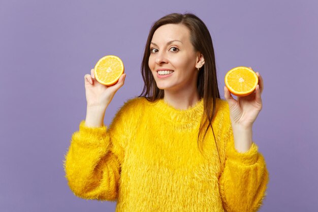 Souriante jeune femme en pull de fourrure tenant dans les mains des moitiés de fruits orange mûrs frais isolés sur fond pastel violet en studio. Les gens vivent un style de vie vivant et se détendent. Maquette de l'espace de copie.