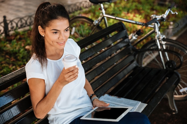 Souriante jeune femme reposant sur le banc avec une tasse de café dans le parc de la ville et tenant la tablette