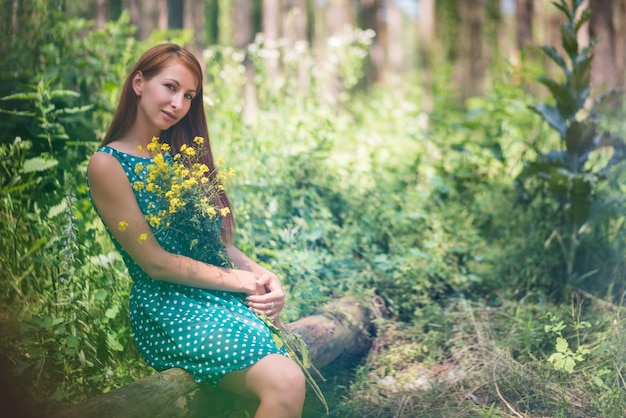 Souriante jeune femme en robe verte assise sur le journal et tenant des fleurs jaunes dans la forêt le jour d'été ensoleillé.