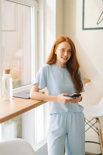 Souriante jeune femme séduisante utilisant un téléphone, tapant des messages en ligne debout près de la fenêtre du café.