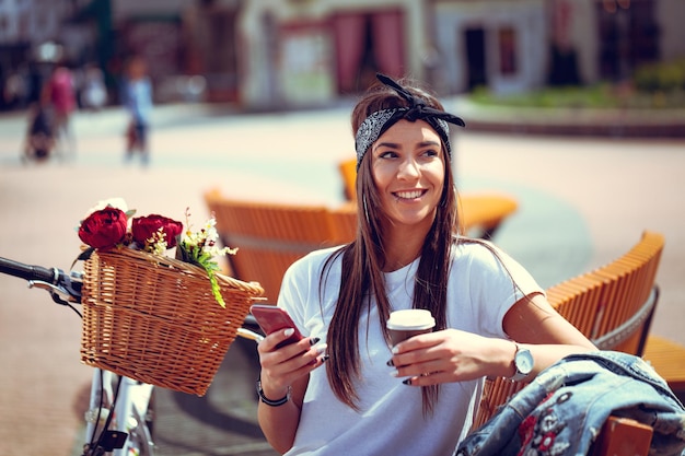 Souriante jeune femme avec smartphone, prenant une pause-café et assise sur un banc par une journée ensoleillée, à côté du vélo avec panier de fleurs.