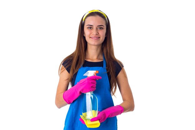 Souriante jeune femme en T-shirt bleu et tablier avec des gants roses tenant des choses de nettoyage dans un lavabo