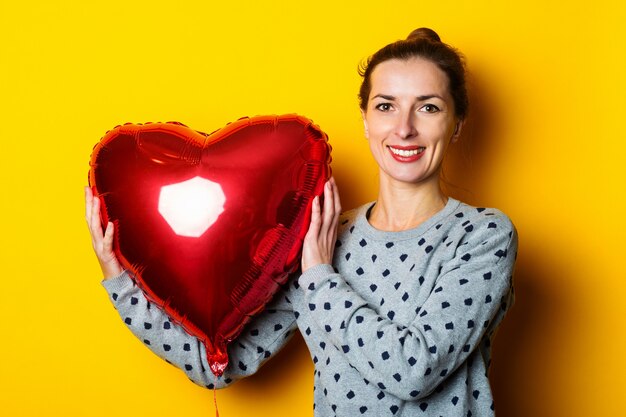 Souriante jeune femme tenant un ballon à air en forme de coeur rouge sur fond jaune.