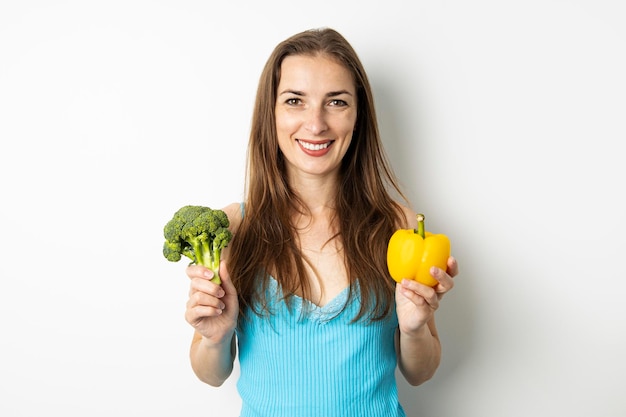 Souriante jeune femme tenant du brocoli et du paprika vert sur fond blanc