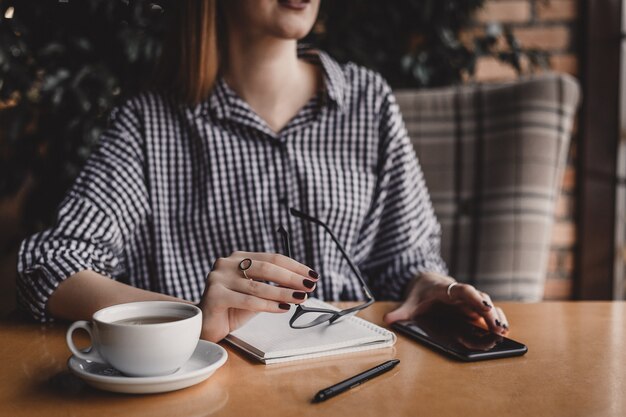 Souriante jeune femme tenant des lunettes tout en étant assis à table avec une tasse de café au café