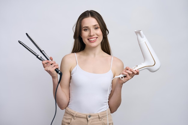 Souriante jeune femme tenant des outils de coiffure lisseur et sèche-cheveux isolé sur fond blanc