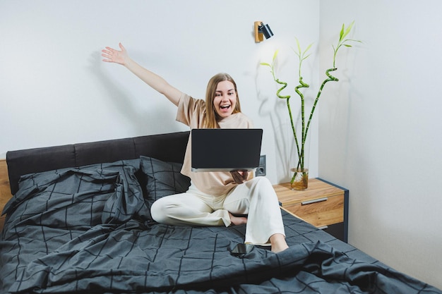 Souriante jeune femme en tenue décontractée souriante et travaillant à distance assise sur le lit avec un ordinateur portable pendant le travail à distance Travail indépendant Travail à domicile