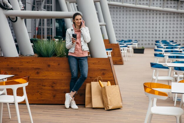 Souriante jeune femme utilise un smartphone pour prendre un taxi après le shopping et la tenue d'une tasse de café, un sac en papier d'épicerie avec une longue baguette de pain blanc