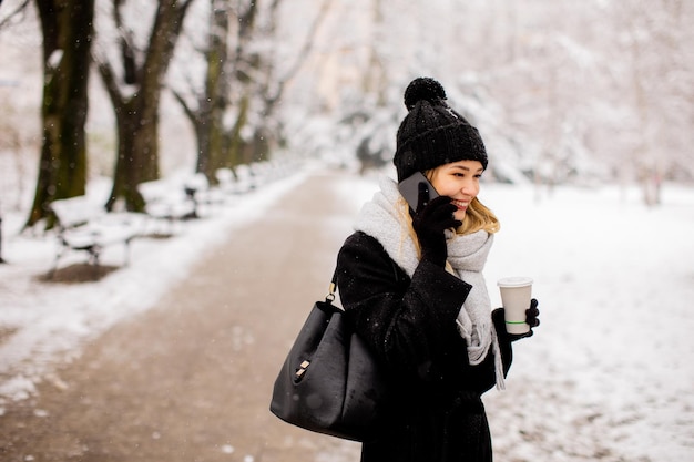 Souriante jeune femme en vêtements confortables utilisant un téléphone portable et tenant une tasse de café le jour de l'hiver