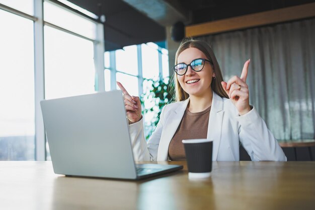 Souriante jeune femme en vêtements décontractés et lunettes portant une veste blanche et riant assis à une table avec un ordinateur portable tout en travaillant