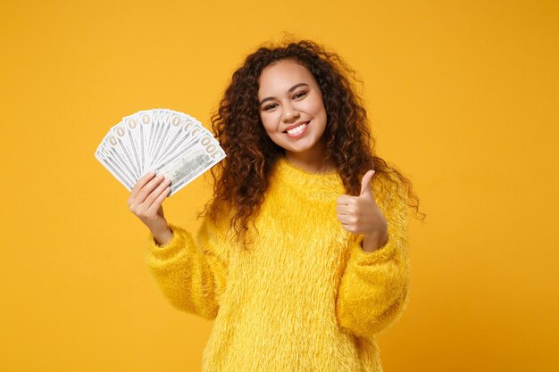 Souriante jeune fille afro-américaine en pull de fourrure posant isolé sur fond jaune. Concept de style de vie des gens. Maquette de l'espace de copie. Tenant un ventilateur d'argent liquide en billets de banque en dollars, montrant le pouce vers le haut.