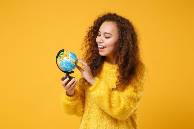 Photo souriante jeune fille afro-américaine en pull de fourrure posant isolé sur fond de mur orange jaune portrait en studio. concept de mode de vie des émotions sincères des gens. maquette de l'espace de copie. tenir le globe terrestre.