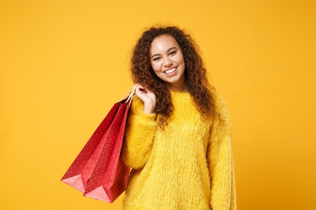 Souriante jeune fille afro-américaine en pull de fourrure posant isolé sur fond orange jaune en studio. Concept de style de vie des gens. Maquette de l'espace de copie. Tenez le sac d'emballage avec les achats après le shopping.