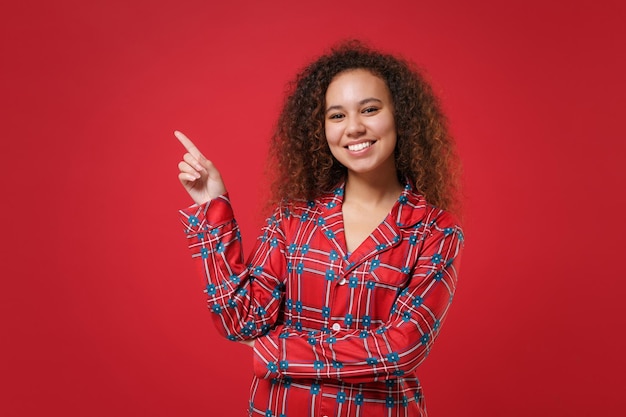 Souriante jeune fille afro-américaine en pyjama homewear posant tout en se reposant à la maison isolée sur fond rouge. Détendez-vous le concept de mode de vie de bonne humeur. Maquette de l'espace de copie. Pointant l'index vers le haut de côté.