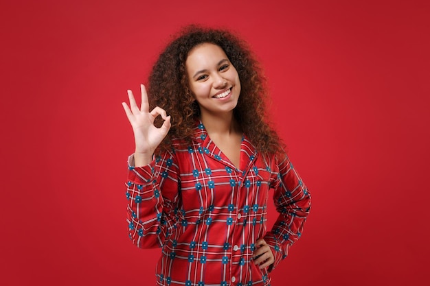 Souriante jeune fille afro-américaine en pyjama homewear posant tout en se reposant à la maison isolée sur fond rouge en studio. Détendez-vous le concept de mode de vie de bonne humeur. Maquette de l'espace de copie. Affichage du geste OK.