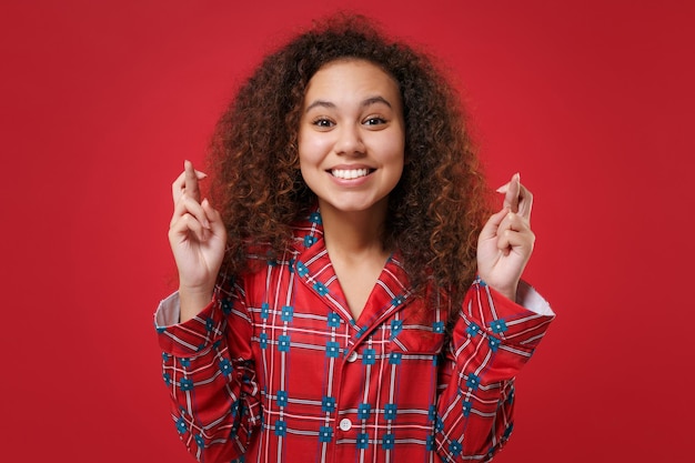 Souriante jeune fille afro-américaine en pyjama homewear reposant à la maison isolée sur fond rouge. Détendez-vous le concept de mode de vie de bonne humeur. Attendre un moment spécial, croiser les doigts, faire un vœu.