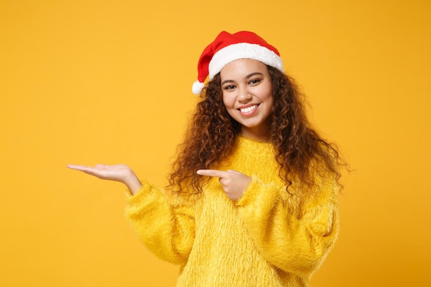 Souriante jeune fille afro-américaine Santa en chapeau de Noël isolé sur fond de mur jaune. Bonne année 2020 concept de vacances de célébration. Maquette de l'espace de copie. Index pointé, main de côté.