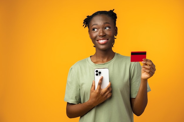 Souriante jeune fille afro détient un téléphone intelligent et une carte de crédit en studio jaune