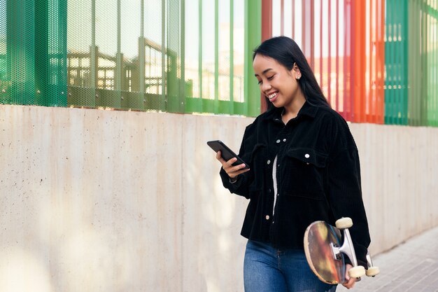 Souriante jeune fille asiatique en train de taper sur le téléphone portable en marchant dans la rue avec sa planche à roulettes à la main