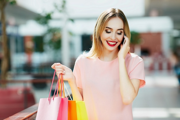 Souriante jeune fille aux cheveux brun clair et lèvres rouges debout avec des sacs colorés, tenant un téléphone mobile, concept de magasinage, portrait, espace de copie.