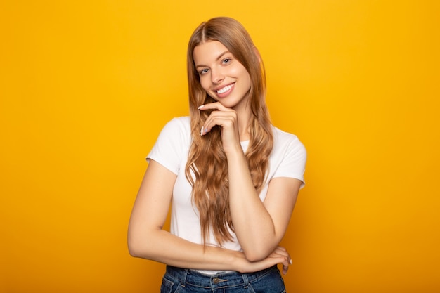 Souriante jeune fille blonde aux cheveux longs touchant le menton isolé sur jaune