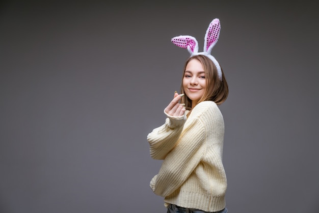 Souriante jeune fille de race blanche, blonde avec des oreilles de lapin, montre un mini coeur avec les doigts isolés sur mur gris