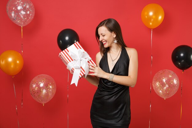 Souriante jeune fille en robe noire célébrant, regardant sur une boîte rouge avec un cadeau, présente dans les mains sur des ballons à air de fond rouge vif. Journée de la femme, bonne année, concept de fête de vacances de maquette d'anniversaire.