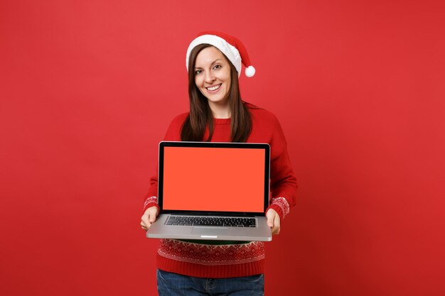 Souriante jeune fille de Santa au chapeau de Noël tenant un ordinateur portable avec un écran vide vierge isolé sur fond de mur rouge. Bonne année 2019 concept de fête de vacances célébration. Maquette de l'espace de copie.
