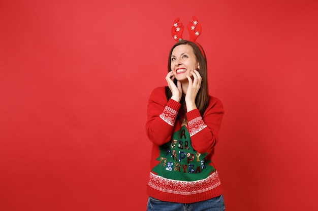 Souriante jeune fille de Santa en pull, cornes de cerf décoratives amusantes en levant, mettant les mains sur le visage isolé sur fond rouge. Bonne année 2019 concept de fête de vacances célébration. Maquette de l'espace de copie.