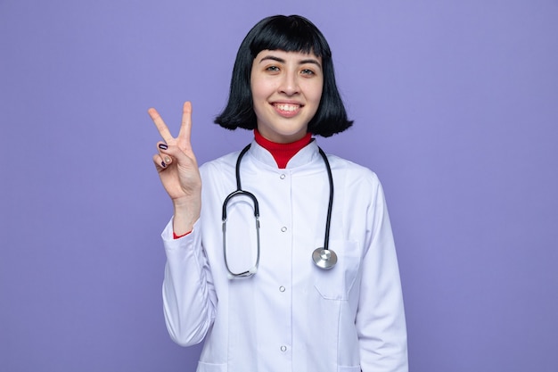 Souriante jeune jolie fille caucasienne en uniforme de médecin avec stéthoscope gesticulant signe de victoire avec la main