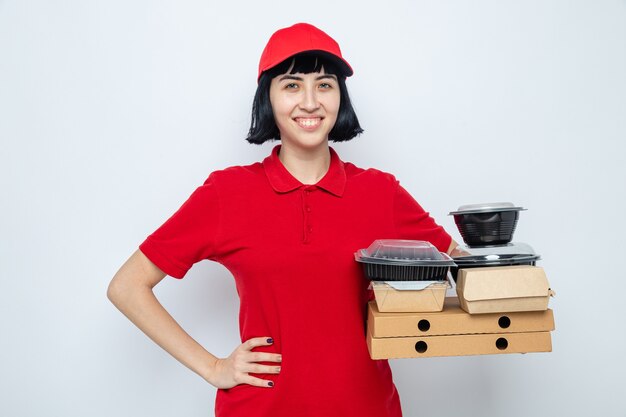Souriante jeune livreuse caucasienne mettant la main sur sa taille et tenant des récipients alimentaires et des emballages sur des boîtes à pizza