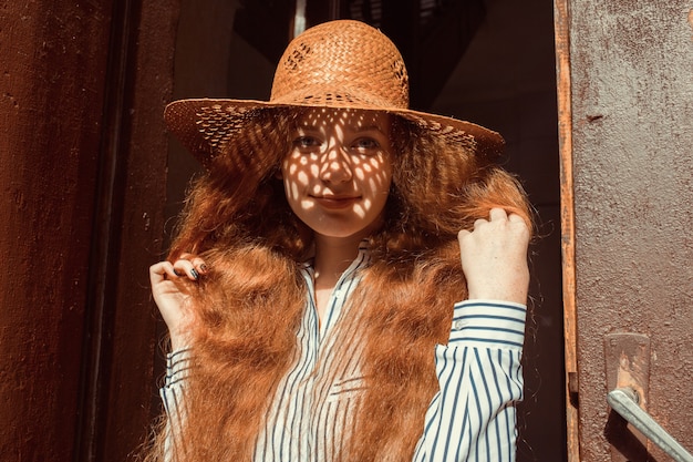 Souriante jeune mannequin aux cheveux rouges au chapeau de paille avec une ombre sur son visage