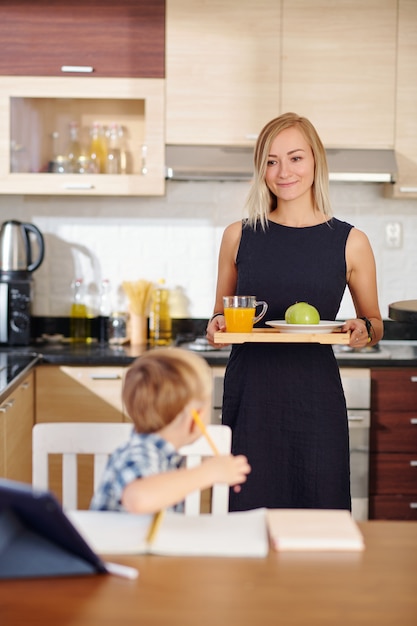 Souriante jeune mère apportant un plateau avec un déjeuner sain à son enfant qui étudie à la table de la cuisine