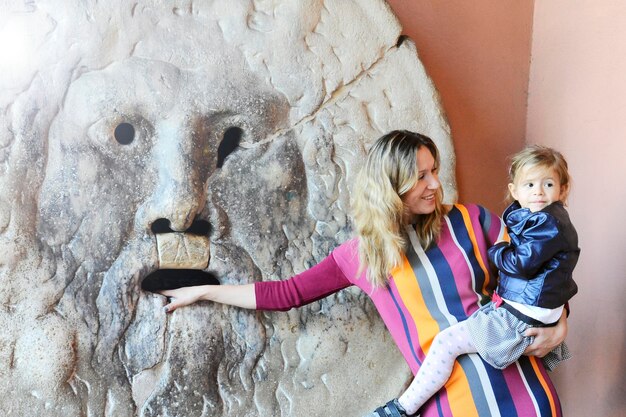 Souriante jeune mère et fille touristes à la bouche de la vérité s'amusant Robe colorée pour mère et veste en cuir bleu pour fille Situation amusante Rome Italie