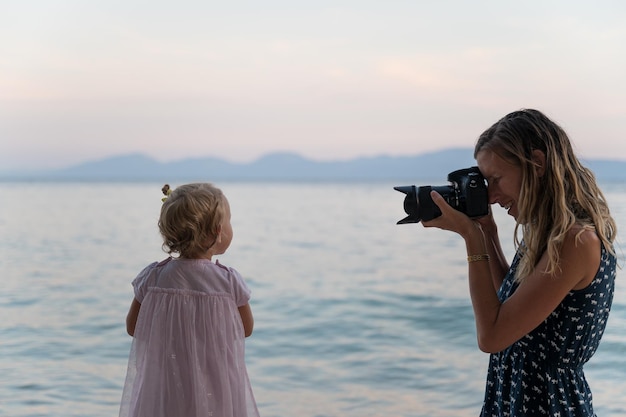 Souriante jeune mère prenant une photo de sa petite fille debout sur la plage juste à côté de la mer du soir