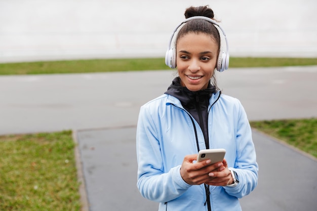 souriante jeune sportive africaine en bonne santé écoutant de la musique avec des écouteurs sans fil s'exerçant au stade, tenant un téléphone portable