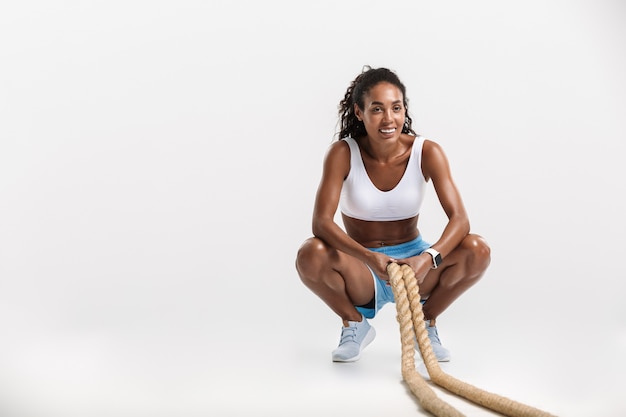 Souriante jeune sportive africaine faisant des exercices avec une corde isolée sur un mur blanc