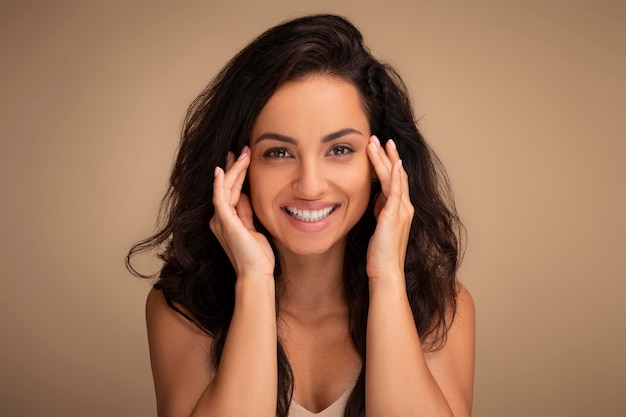 Souriante jolie brune aux cheveux longs jeune femme touchant les tempes de la peau du visage