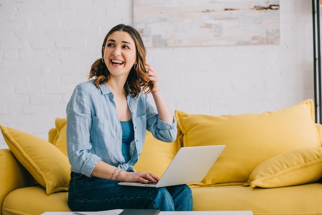 Souriante jolie femme assise sur un canapé jaune avec ordinateur portable