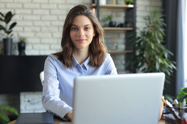 Souriante jolie femme assise à table, regardant la caméra. Heureux entrepreneur lisant un message électronique avec de bonnes nouvelles, discutant avec des clients en ligne.