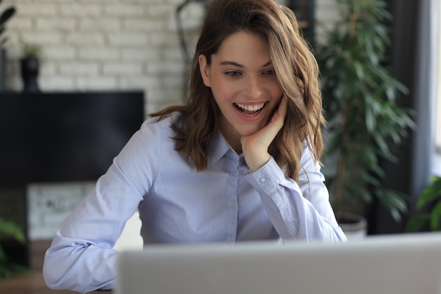 Photo souriante jolie femme assise à table, regardant l'écran d'un ordinateur portable. heureux entrepreneur lisant un message électronique avec de bonnes nouvelles, discutant avec des clients en ligne.