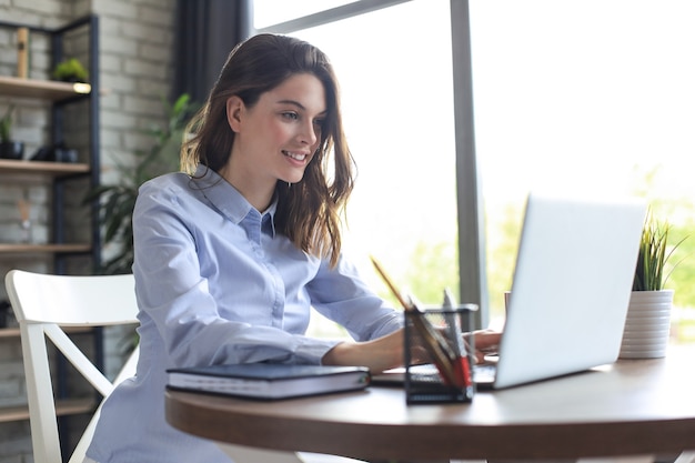 Souriante jolie femme assise à table, regardant l'écran d'un ordinateur portable. Heureux entrepreneur lisant un message électronique avec de bonnes nouvelles, discutant avec des clients en ligne.