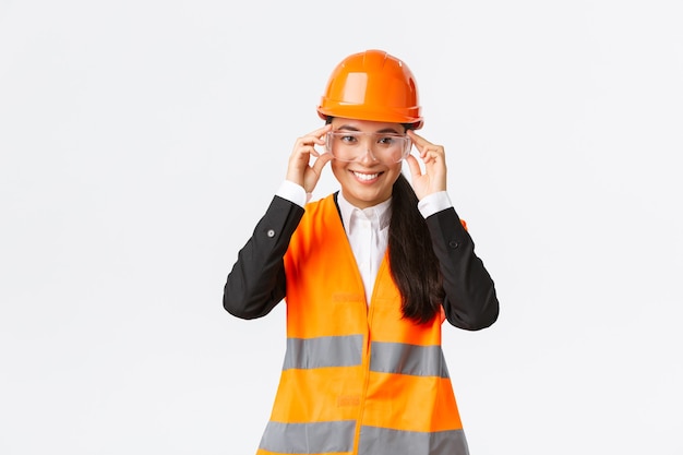 Souriante jolie femme ingénieur asiatique, responsable de la construction portant un casque de sécurité et des lunettes suivant le protocole avant d'entrer dans la zone de construction ou l'entreprise, debout sur fond blanc