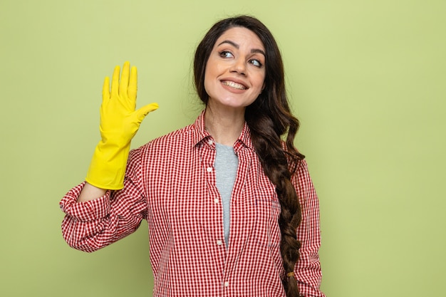 Souriante jolie femme de ménage caucasienne avec des gants en caoutchouc gardant la main ouverte et regardant de côté