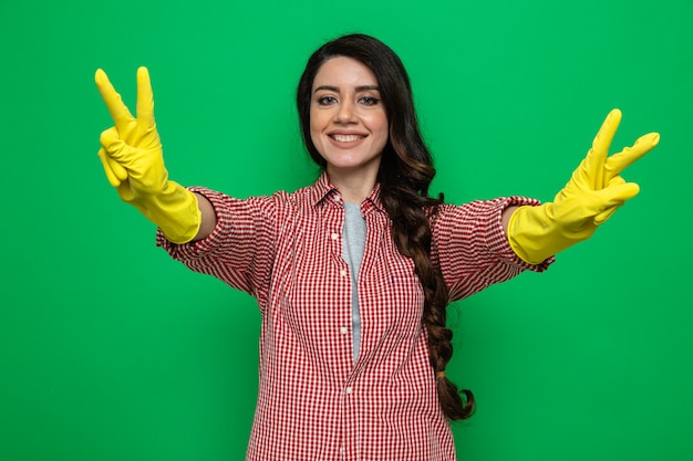 Souriante jolie femme de ménage caucasienne avec des gants en caoutchouc gesticulant le signe de la victoire à deux mains