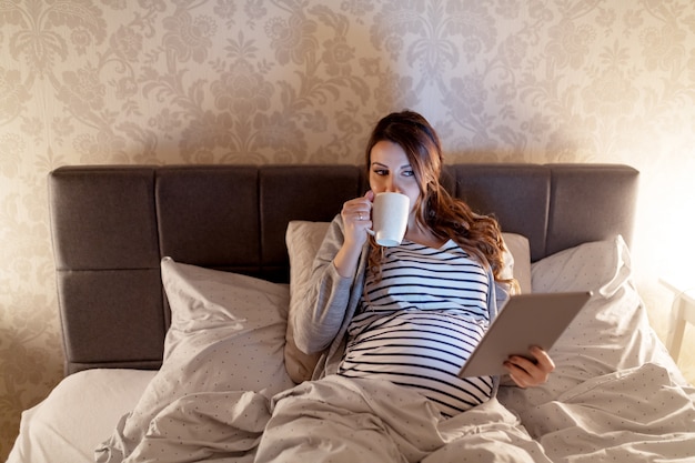 Souriante Jolie Femme De Race Blanche Enceinte Aux Longs Cheveux Bruns Couché Dans Le Lit, Boire Du Thé Et Lire Sur Les Bébés Sur Tablette. Soirée.