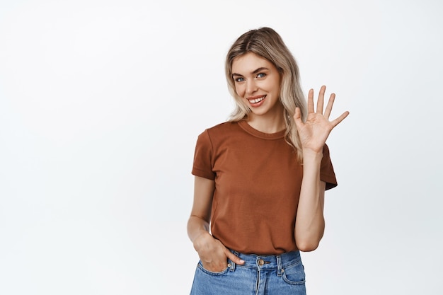 Souriante Jolie Fille Agitant La Main Et Dit Bonjour, Vous Saluant Avec Un Visage Coquettement Heureux, Debout Sur Blanc En Tenue Décontractée.