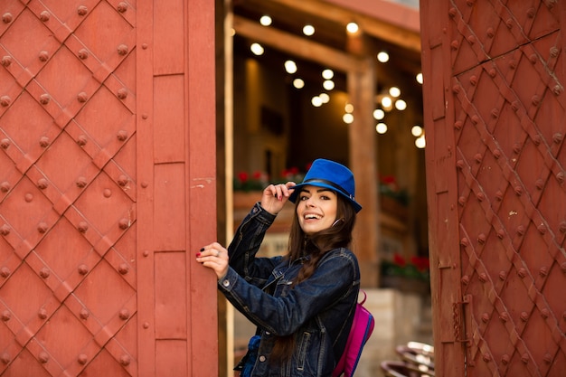 Souriante jolie fille au chapeau bleu près du vieil immeuble avec des portes rouges antiques. Modèle féminin pose