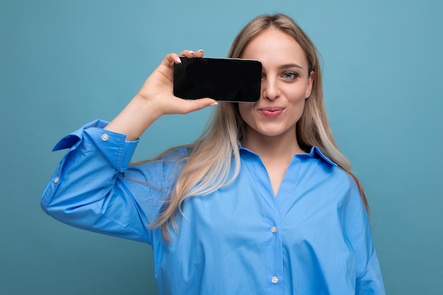 Souriante jolie fille blonde tenant un smartphone avec une maquette d'écran vierge pour une page Web sur bleu