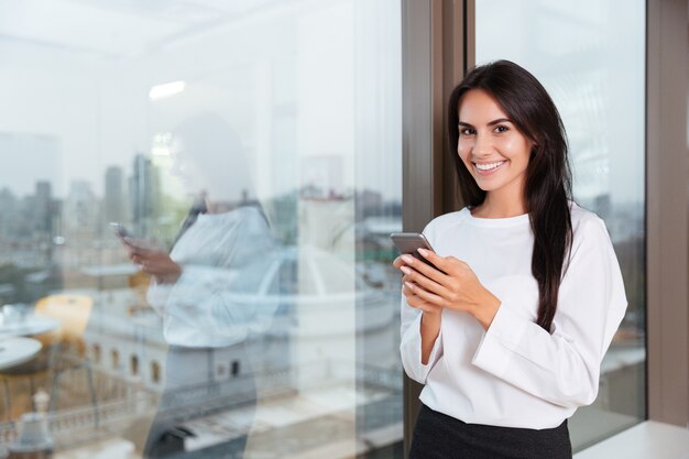 Souriante jolie jeune femme d'affaires utilisant un smartphone près de la fenêtre au bureau